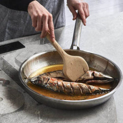 Person cooking fish in a pan with a wooden spatula on a kitchen counter.