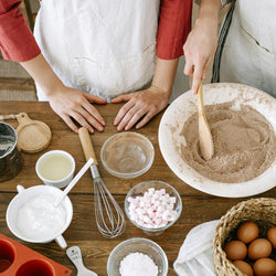 Two people preparing ingredients on a wooden table with various kitchen utensils and items.